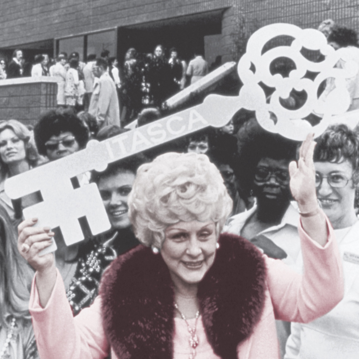 Mary Kay Ash, die Firmengr&uuml;nderin des Direktvertriebsunternehmens Mary Kay, ist auf einem alten Foto im Vordergrund in einem rosa Kost&uuml;m zu sehen. Sie h&auml;lt mit beiden H&auml;nden einen gro&szlig;en Schl&uuml;ssel in die H&ouml;he. Hinter ihr sind viele weitere Menschen zu sehen.